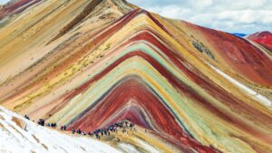 A wide, stunning view of Vinicunca, the Rainbow Mountain in Peru, showing its famous colorful mineral stripes with a crowd of travelers at the viewpoint – Qosqo Expeditions