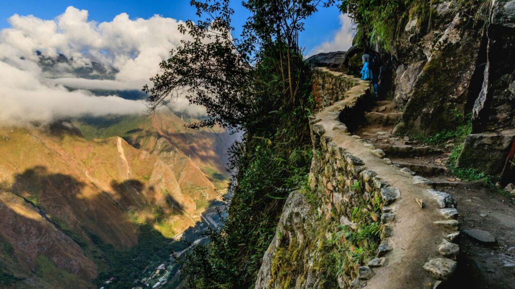 A hiker on the narrow, stone-paved Inca Trail, carved into a cliffside high in the Andes, with a dramatic view of the cloud-filled valley below – Qosqo Expeditions
