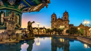 The central fountain in Cusco's Plaza de Armas at dusk, with the warm, illuminated facade of the historic Cusco Cathedral in the background – Qosqo Expeditions