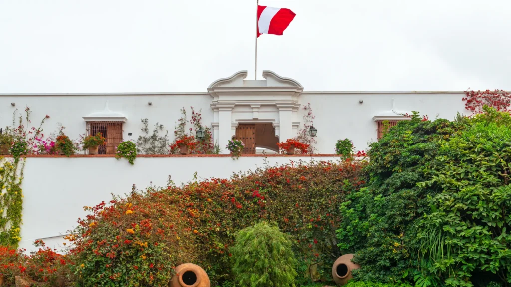 The beautiful white colonial facade of the Larco Museum in Lima, surrounded by vibrant flowering gardens, with the Peruvian flag flying above. – Qosqo Expeditions