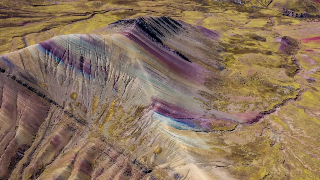 An aerial view of the colorful mineral stripes of Rainbow Mountain (Vinicunca) in Peru, showing the vibrant geological formation set within the vast Andean landscape. – Qosqo Expeditions
