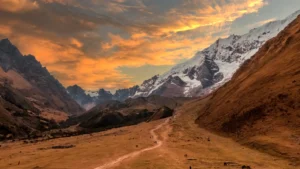 Hikers walking on a winding trail through a vast Andean valley at sunset, with the majestic, snow-covered peak of a sacred mountain in the background. – Qosqo Expeditions