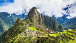 The classic postcard view of the Machu Picchu citadel, with its green terraces and stone ruins, under a partly cloudy sky with Huayna Picchu mountain in the background – Qosqo Expeditions