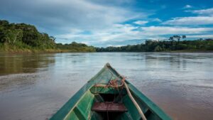 View from the bow of a wooden boat traveling down a wide river through the dense Peruvian Amazon rainforest – Qosqo Expeditions