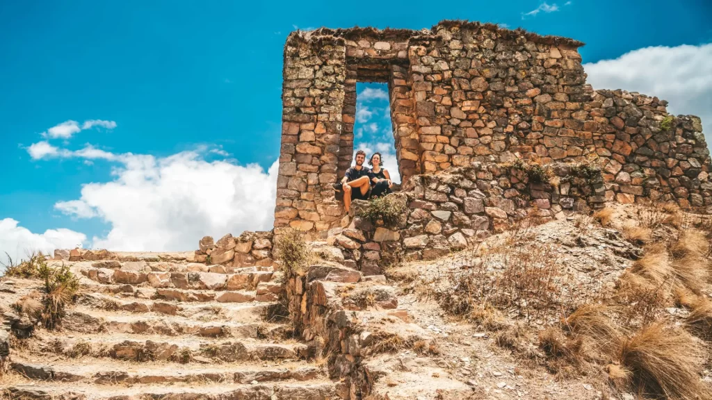 A couple sitting framed within an ancient Inca stone gateway, with a bright blue sky above, representing a moment of discovery and connection during their Inca Trail trek. - Qosqo Expeditions