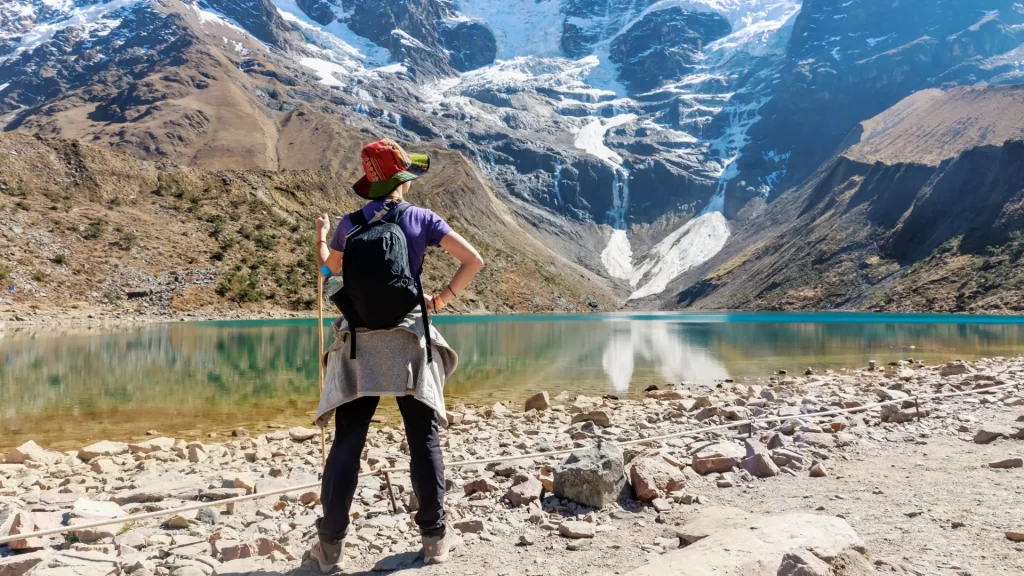 A solo hiker admiring a clear turquoise mountain lake nestled beneath snow-capped peaks, a breathtaking scene ideal for bespoke Peru travel adventures. - Qosqo Expeditions