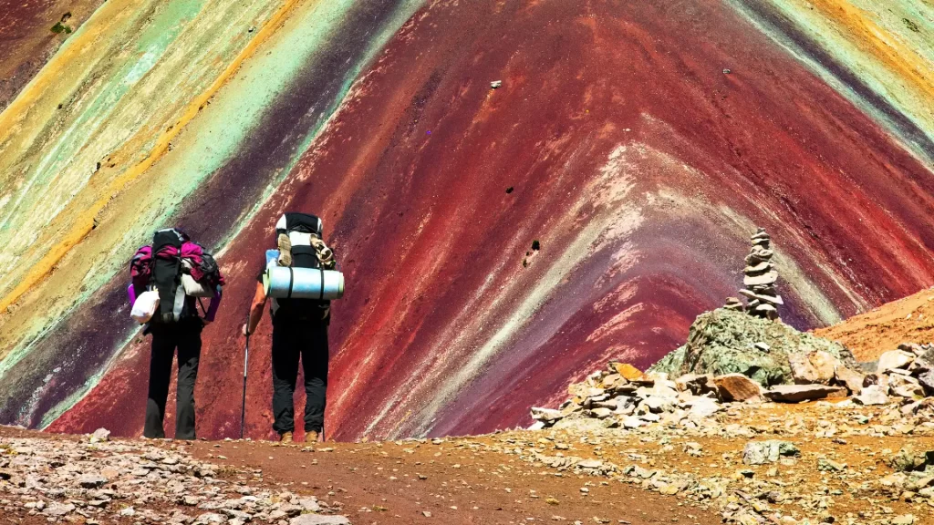 Two hikers with backpacks standing on a rocky path, looking up at the dramatic, richly colored slopes of Rainbow Mountain with a small stone cairn nearby, often led by a local rainbow mountain guide. - Qosqo Expeditions