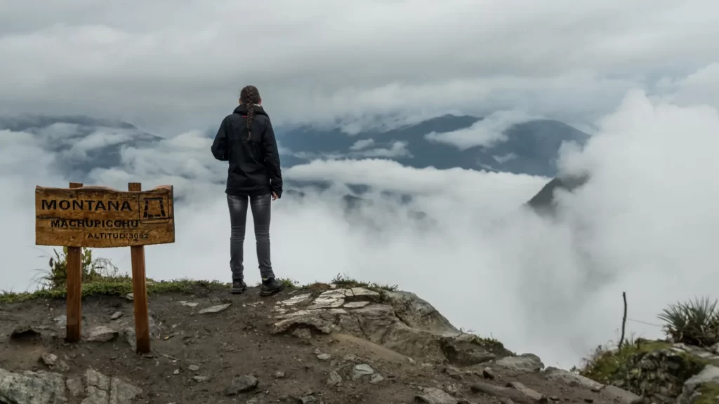 A traveler standing at the sign for Montaña Machu Picchu (Altitude: 3082m), looking out at a breathtaking panoramic view of misty mountains and clouds, offering an exclusive perspective on a short Inca Trail luxury trek. - Qosqo Expeditions