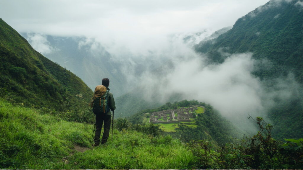 A lone hiker standing on a misty mountain trail, overlooking distant Inca ruins nestled in the valley below, showcasing the unique perspectives on private Machu Picchu tours. - Qosqo Expeditions