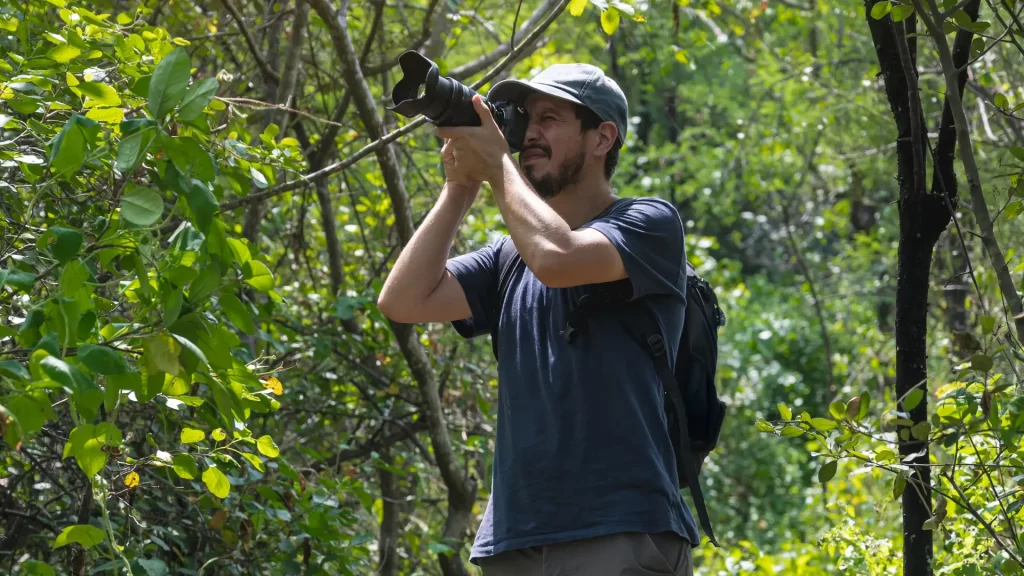 A male photographer in a cap and backpack holding a professional camera, taking pictures in a lush, green forest, indicative of a nature-focused luxury Amazon Andes expedition. - Qosqo Expeditions