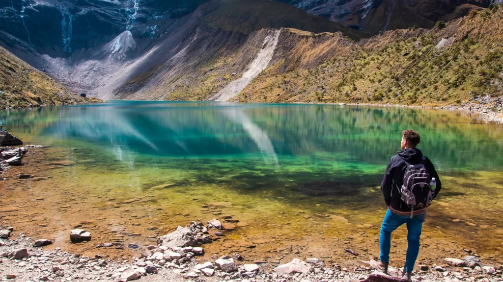 A male hiker with a backpack stands on the rocky shore of Humantay Lake, admiring its crystal-clear turquoise waters reflecting the surrounding mountains, emphasizing the serene experience at Humantay Lake. - Qosqo Expeditions