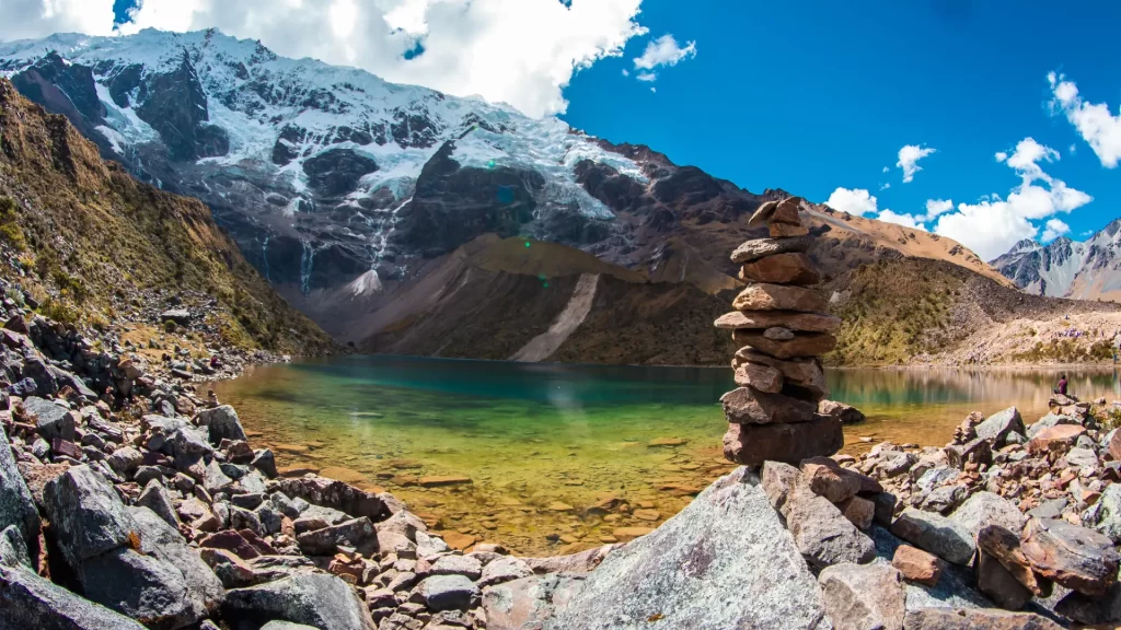A stacked stone cairn (apacheta) in the foreground, with the stunning turquoise waters of Humantay Lake and imposing snow-capped mountains under a blue sky in the background, a common sight near Humantay Lake. - Qosqo Expeditions