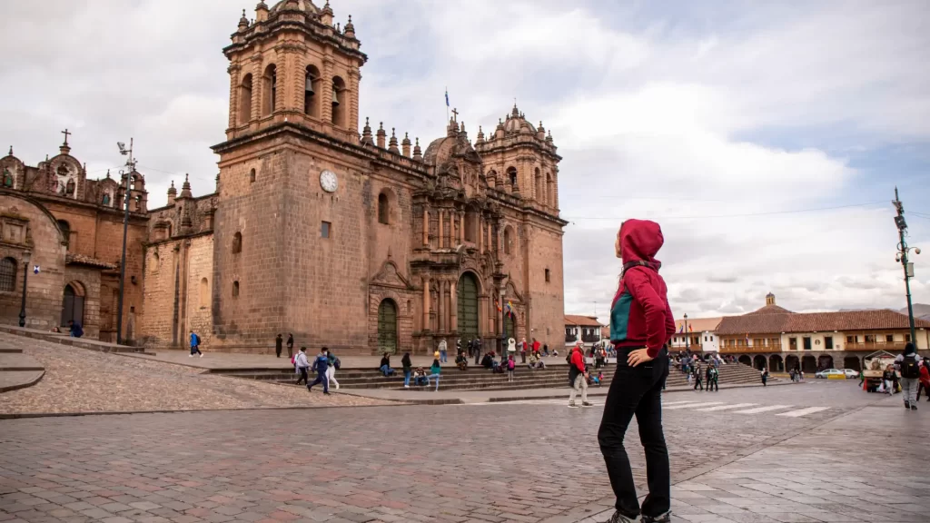 A lone traveler standing in Cusco's Plaza de Armas, looking towards the grand Cusco Cathedral under a cloudy sky, a central feature of any Peru itinerary focused on culture and history. - Qosqo Expeditions