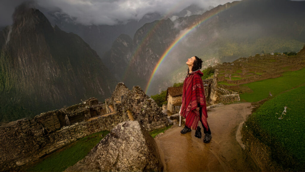 A female tourist in a red poncho gazing up at a vibrant rainbow arching over the ancient Inca city of Machu Picchu, a truly magical moment experienced on private Machu Picchu tours. - Qosqo Expeditions