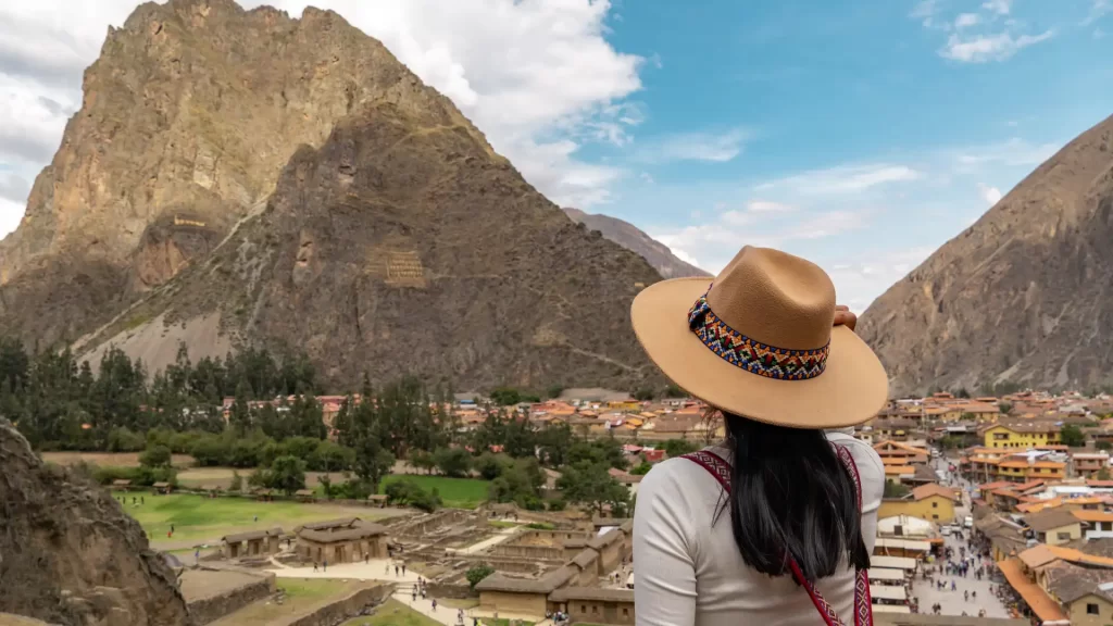 A traveler with a traditional hat, viewed from behind, gazing at the impressive Inca ruins and town of Ollantaytambo nestled in the Sacred Valley, a key stop on any comprehensive Peru itinerary. - Qosqo Expeditions