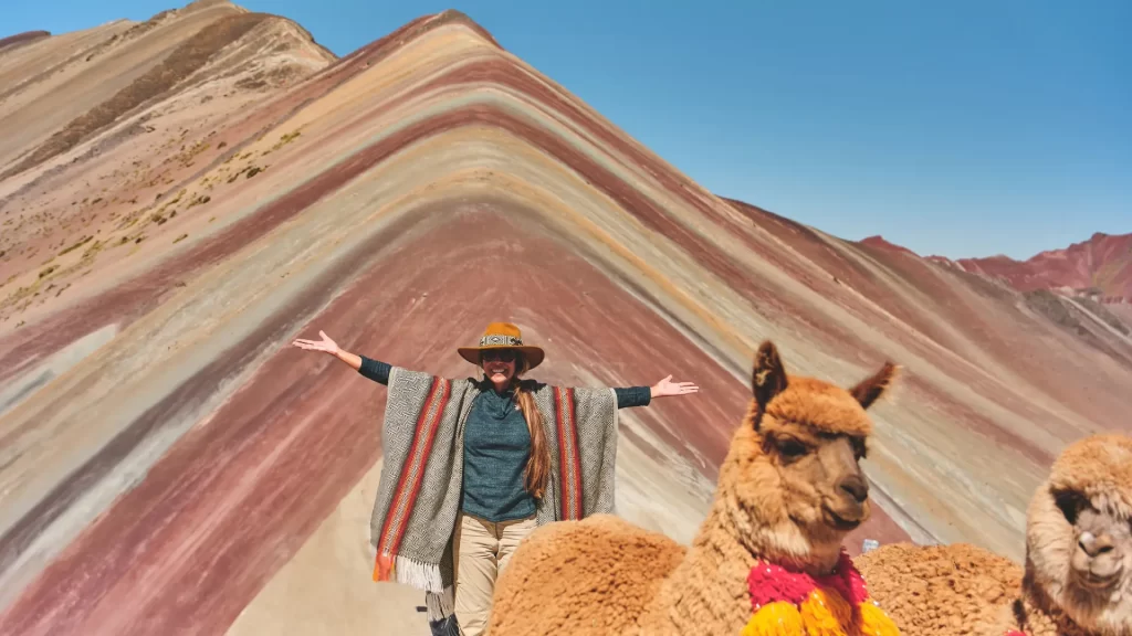 A joyful tourist with arms outstretched, posing with llamas in front of the stunning colorful slopes of Rainbow Mountain, a memorable experience enhanced by a rainbow mountain guide. - Qosqo Expeditions