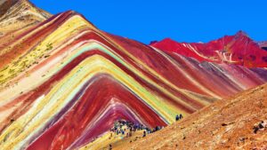 A stunning wide shot of Vinicunca, the Rainbow Mountain, showing its vibrant mineral stripes with a crowd of hikers on the ridge under a blue sky – Qosqo Expeditions
