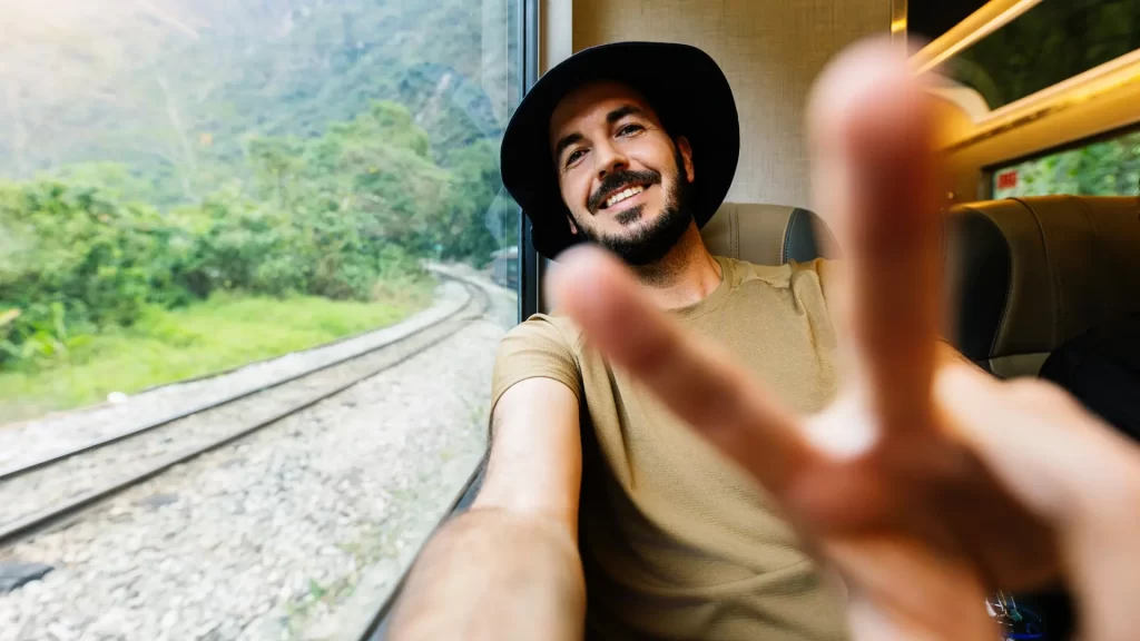 A happy male traveler in a hat taking a selfie inside a train with lush green scenery and train tracks passing by, a comfortable segment of bespoke Peru travel. - Qosqo Expeditions