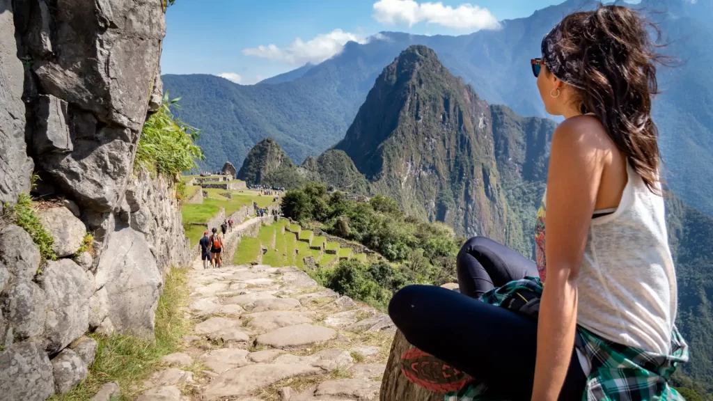 A female traveler sitting on a stone wall, gazing at the panoramic view of Machu Picchu citadel and Huayna Picchu mountain, a stunning reward from an Inca Trail trek. - Qosqo Expeditions