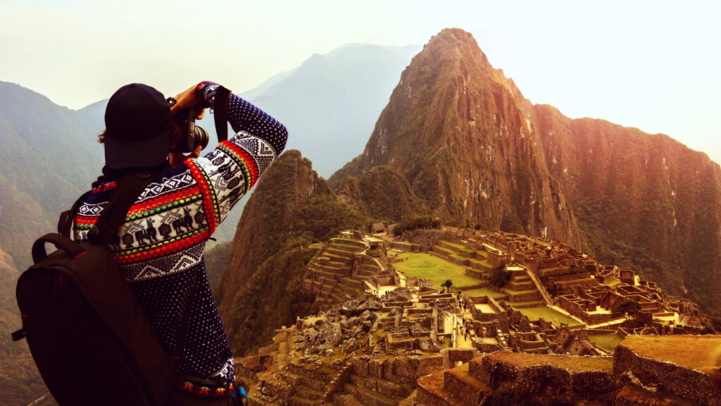 A traveler from behind, wearing a traditional Peruvian sweater, photographing the ancient Inca citadel of Machu Picchu at sunset, perfect for private Machu Picchu tours. - Qosqo Expeditions