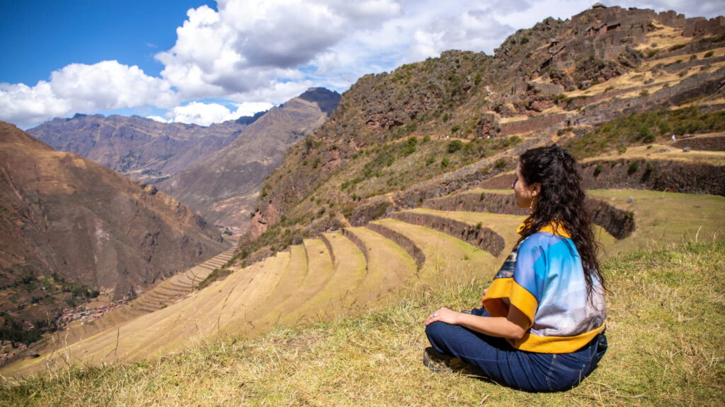A woman sitting peacefully on a grassy hill, looking out over the magnificent ancient Inca agricultural terraces of the Pisac archaeological site in the Sacred Valley of Peru – Qosqo Expeditions