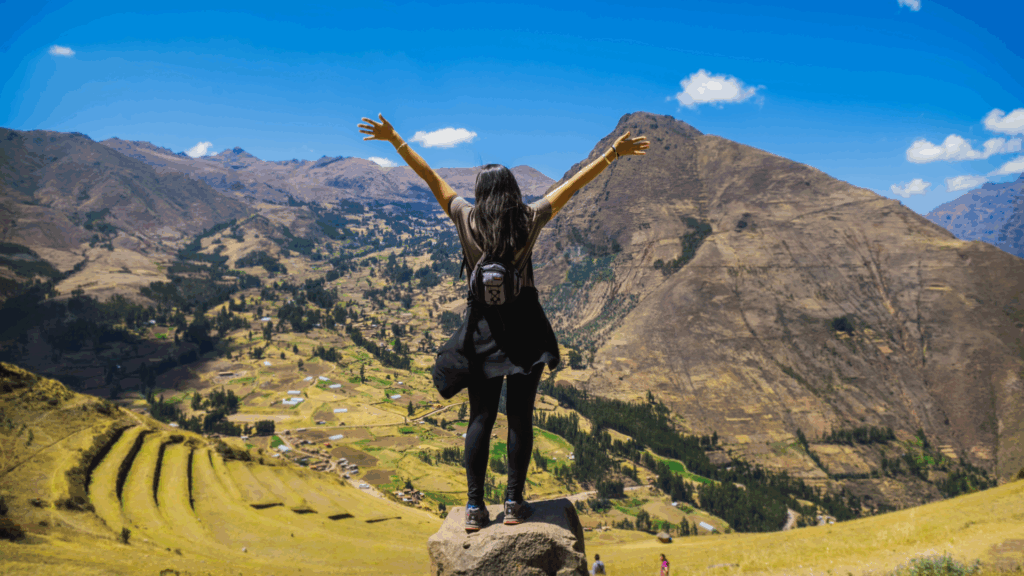 A woman viewed from behind with arms outstretched in joy, standing on a viewpoint overlooking the vast green terraces and landscape of the Sacred Valley of the Incas in Peru – Qosqo Expeditions