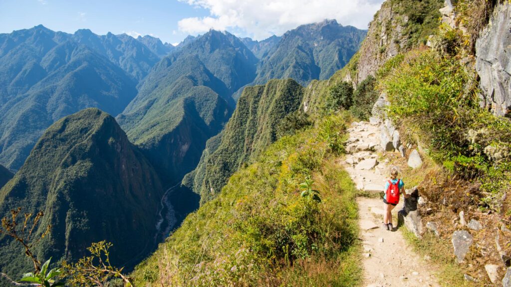 A lone hiker with a red backpack walks along the narrow, stone-paved Inca Trail, carved into a lush green mountainside with panoramic views of the Andes and the valley below – Qosqo Expeditions