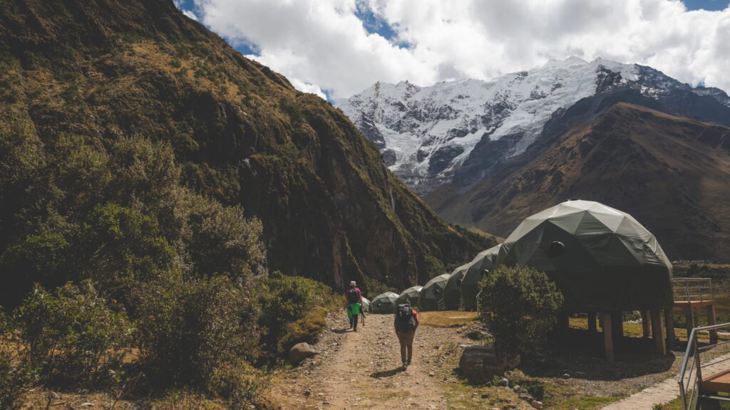 Two hikers walking on a trail towards luxury geodesic sky domes at a campsite in Soraypampa, with the snow-capped Salkantay mountain peak in the background – Qosqo Expeditions