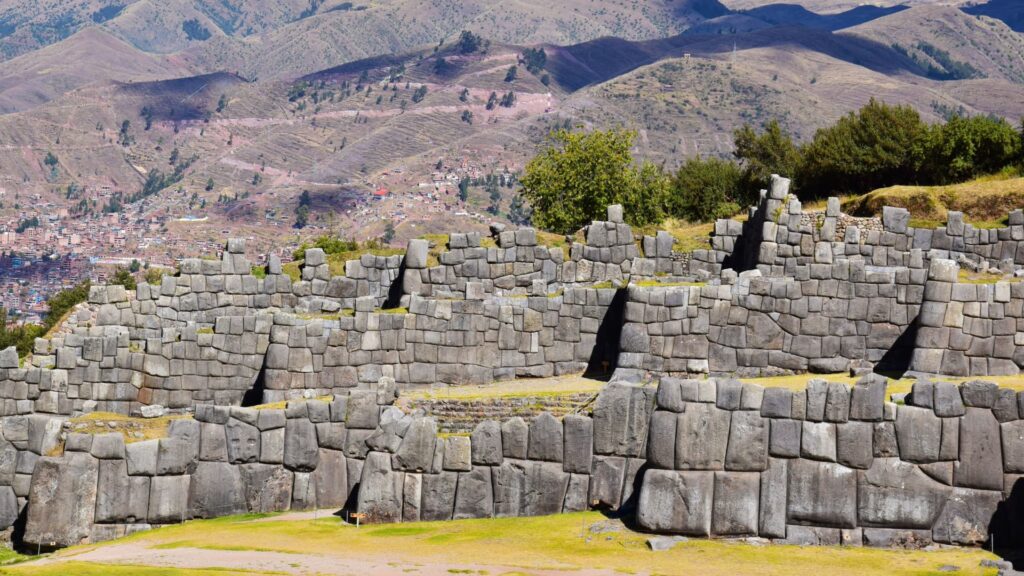 A panoramic view from the Sacsayhuaman Inca fortress, showing the massive stone walls in the foreground with the city of Cusco visible in the valley below and the Andes mountains in the background – Qosqo Expeditions