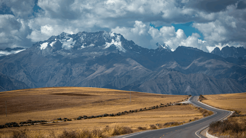 A winding paved road through the golden-brown high plains of the Andes, leading towards massive snow-capped mountains, representing the scenic journey from Arequipa to the Colca Canyon – Qosqo Expeditions
