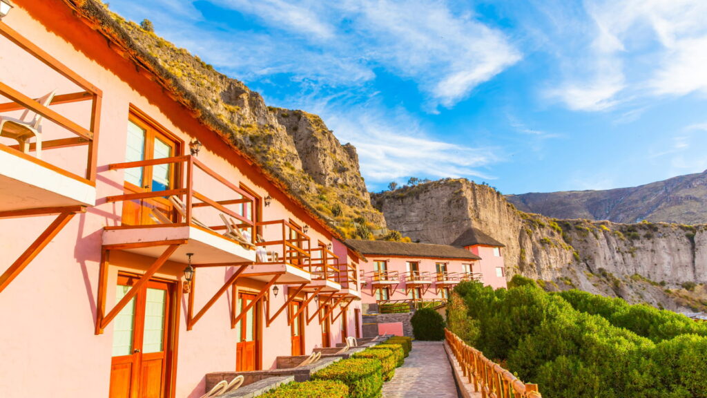 A pink-walled hotel with wooden balconies built into the side of the Colca Canyon, with dramatic cliffs and a bright blue sky, showcasing a unique lodge in the Arequipa region of Peru – Qosqo Expeditions