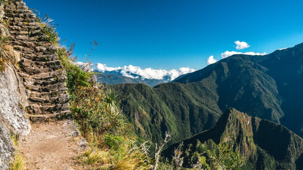 View of ancient Inca stone steps alongside a dirt path, with lush green mountains and a clear blue sky in the background, depicting the iconic Inca Trail – Qosqo Expeditions