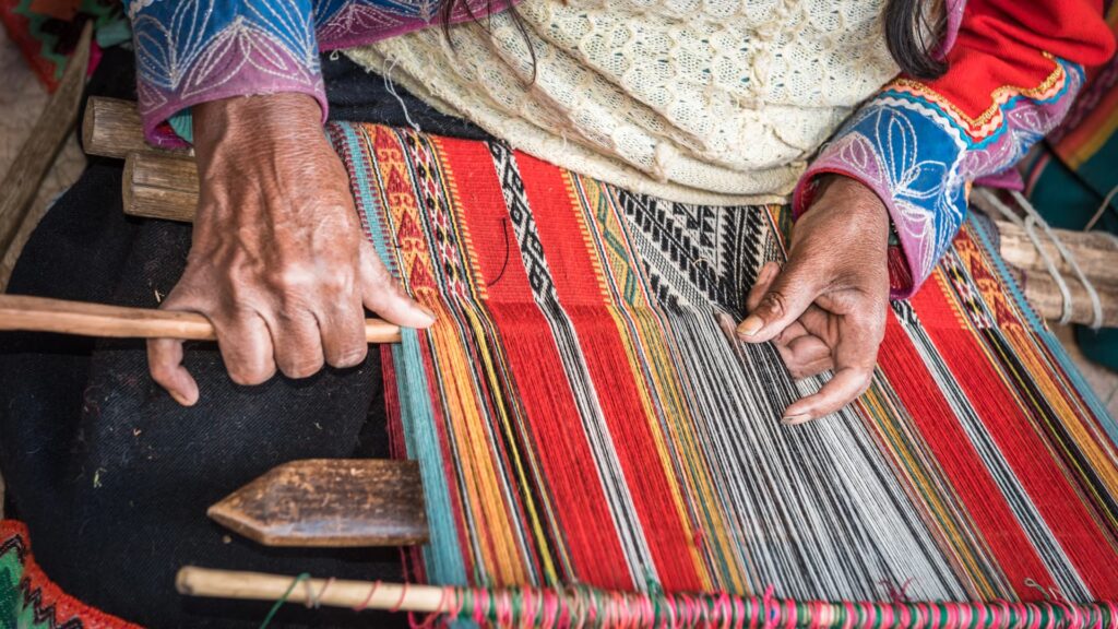 Close-up of an indigenous Andean woman's hands expertly weaving colorful, traditional Peruvian textiles on a backstrap loom, showcasing ancient artisanal techniques in the Sacred Valley – Qosqo Expeditions