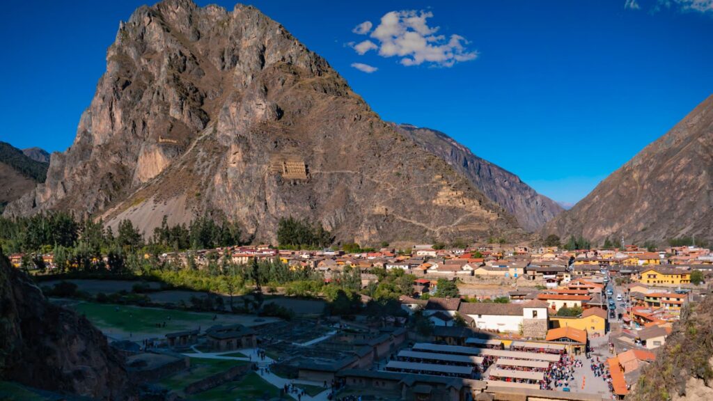 Wide view of the Inca town and fortress of Ollantaytambo, with the ancient Pinkuylluna granaries carved into the mountain across the valley, under a clear blue sky – Qosqo Expeditions