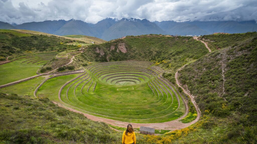 Wide shot of the circular Inca agricultural terraces of Moray, with a traveler in a yellow jacket observing the site, surrounded by green fields and distant mountains in the Sacred Valley, Peru – Qosqo Expeditions
