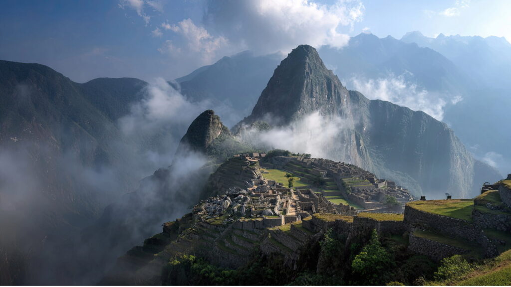 Panoramic view of the ancient Inca city of Machu Picchu emerging from the morning mist and clouds, with Huayna Picchu mountain in the background, under a dramatic sky, conveying a sense of peace and wonder – Qosqo Expeditions Description (EN): Experience the profound magic of Machu Picchu as it awakens. This breathtaking image captures the iconic Inca citadel enveloped in soft morning clouds, offering a serene and mystical panorama that invites contemplation and discovery, a hallmark of our exclusive journeys.