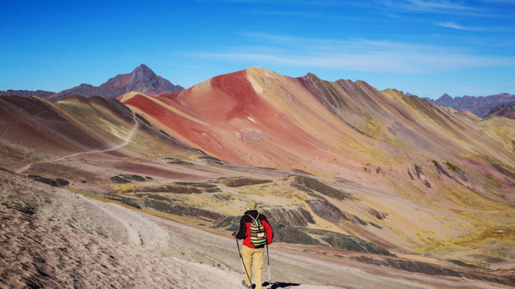 A solitary hiker walks along a path admiring the panoramic views of multiple colorful Palcoyo Rainbow Mountains and the vast Andean landscape under a clear blue sky in Peru – Qosqo Expeditions