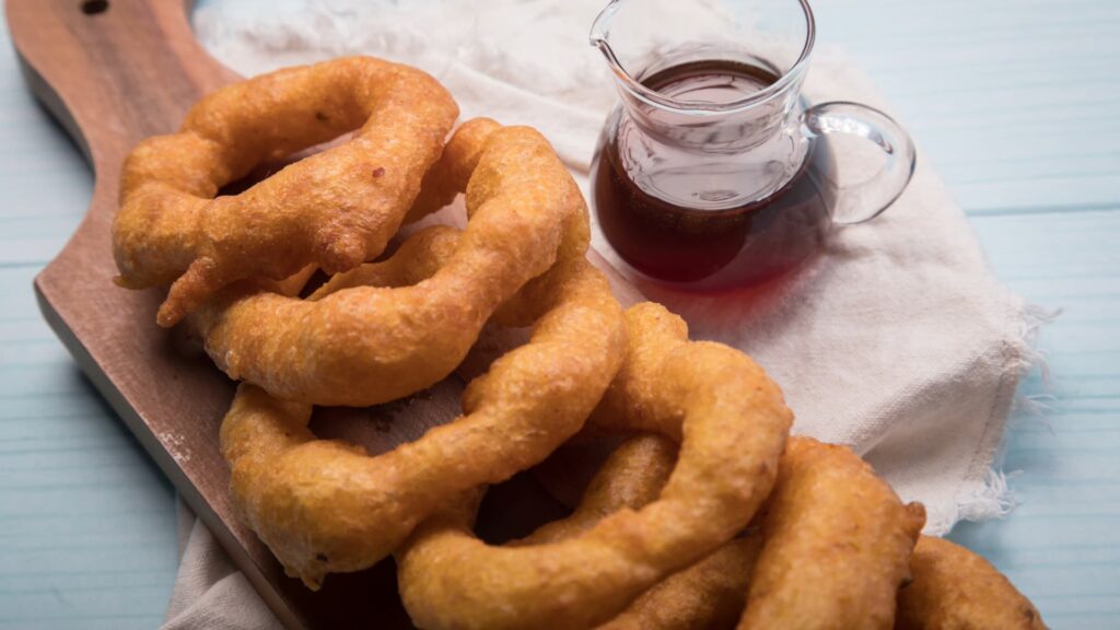A close-up of a pile of golden, freshly fried Peruvian picarones (squash and sweet potato doughnuts) served on a wooden board with a glass pitcher of chancaca syrup – Qosqo Expeditions