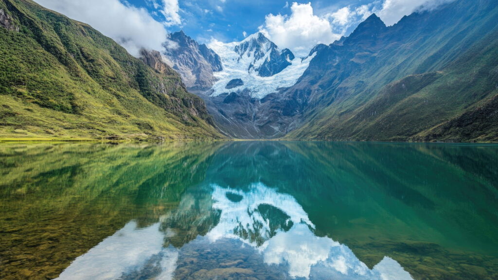The stunning turquoise Humantay Lake with a perfect reflection of the snow-capped Humantay glacier and the Andes mountains under a blue, cloudy sky, a highlight of the Salkantay Trek – Qosqo Expeditions