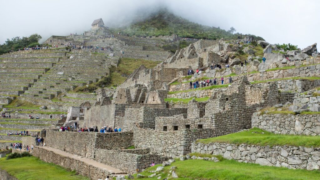 A wide view of the Machu Picchu archaeological site with numerous tourists exploring the ancient Inca stone terraces and ruins on a cloudy day, with Huayna Picchu in the background – Qosqo Expeditions