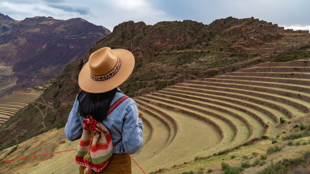 A woman wearing a hat and a traditional Andean bag, viewed from behind, contemplates the vast, curved Inca terraces of Pisac, an ideal setting for a Sacred Valley wellness retreat – Qosqo Expeditions