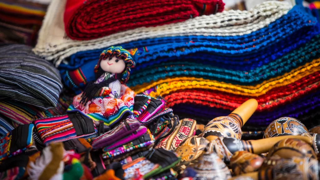 Colorful display of traditional Peruvian textiles, woven belts, carved maracas, and a handmade cloth doll in Andean attire-Qosqo Expeditions