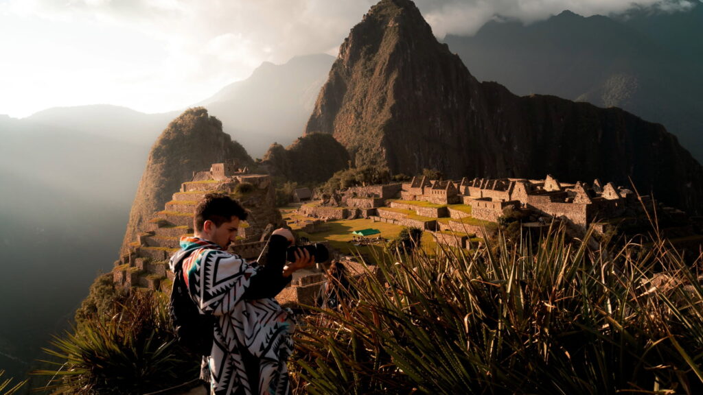 A male photographer in a colorful poncho capturing the ancient citadel of Machu Picchu at sunrise, with Huayna Picchu in the background, a key moment for any Peru photography guide – Qosqo Expeditions