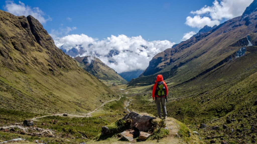 A solo hiker wearing a red jacket stands on a mountain ridge overlooking a stunning green valley surrounded by towering peaks and clouds in the Peruvian Andes.-Qosqo Expeditions