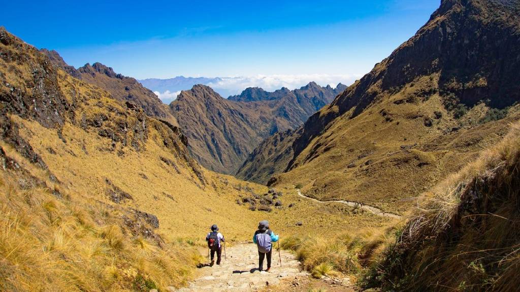 Two tourists walk along the stone trail at the Warmiwañusca Pass under a clear sky, during the best time of year to hike the Inca Trail.