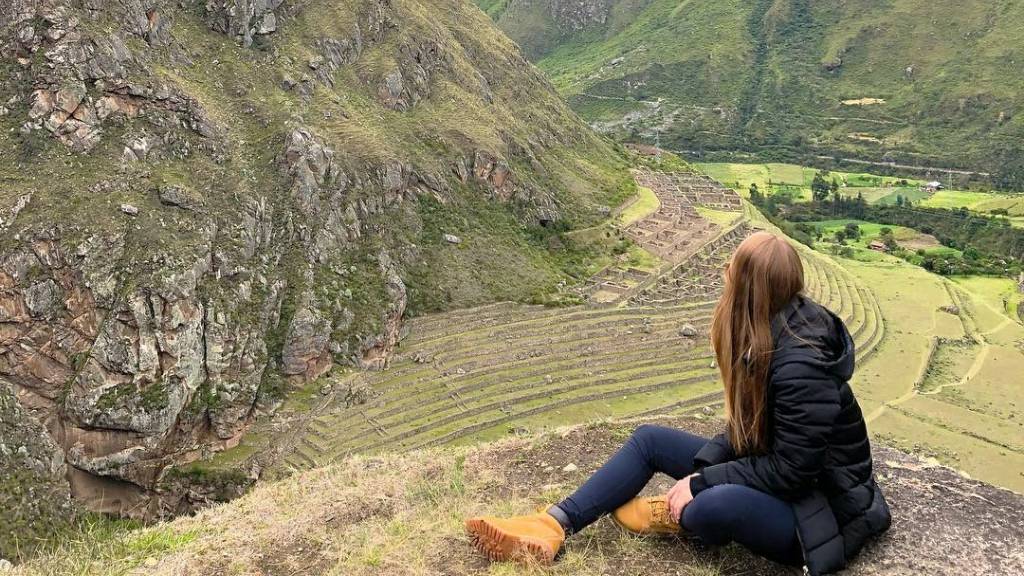 A seated tourist looks out over the Patallaqta archaeological site on a clear day, showcasing the ideal conditions for exploring the Inca Trail during its peak season.