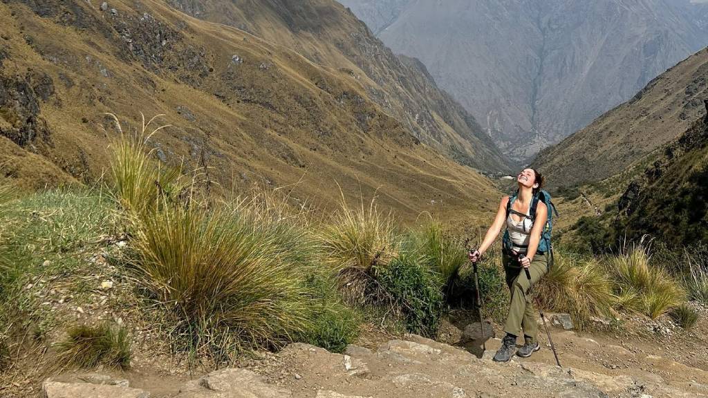 A tourist smiles as she climbs toward the Paso de la Mujer Muerta on the Inca Trail, using trekking poles.