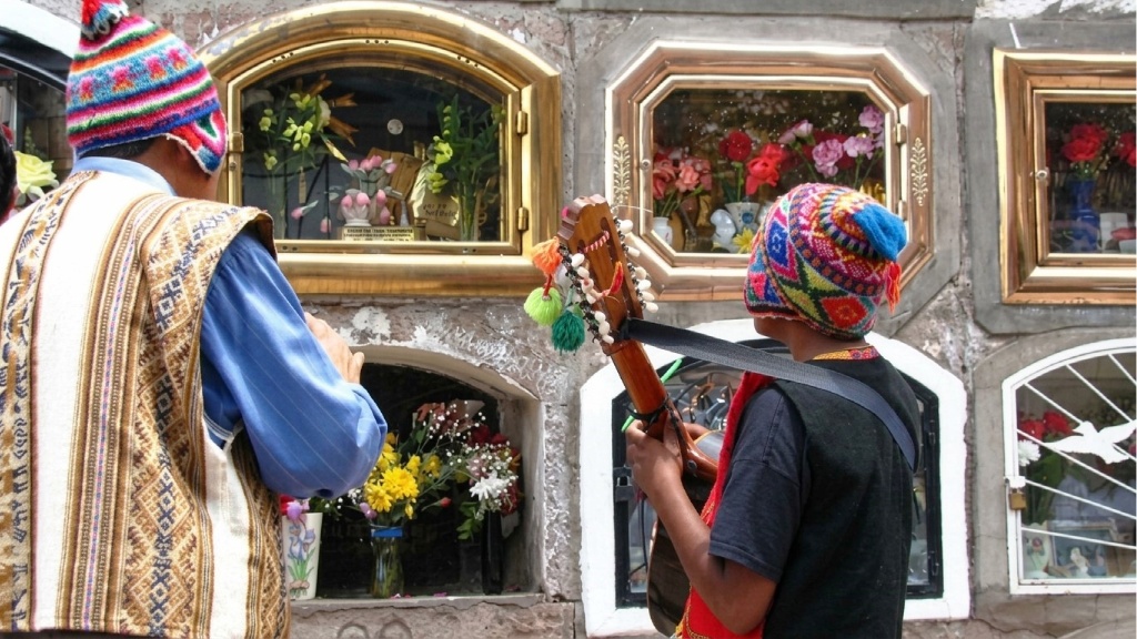 Two musicians wearing traditional Andean chullos and vests playing instruments in front of flower-decorated burial niches during the Day of the Dead in Peru.