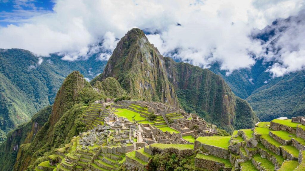 Panoramic view of Machu Picchu with its agricultural terraces and surrounding mountains under a cloudy sky.